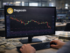 A trader’s hands operate a keyboard while monitoring a Dogecoin candlestick chart on a desktop monitor in a financial office setting.